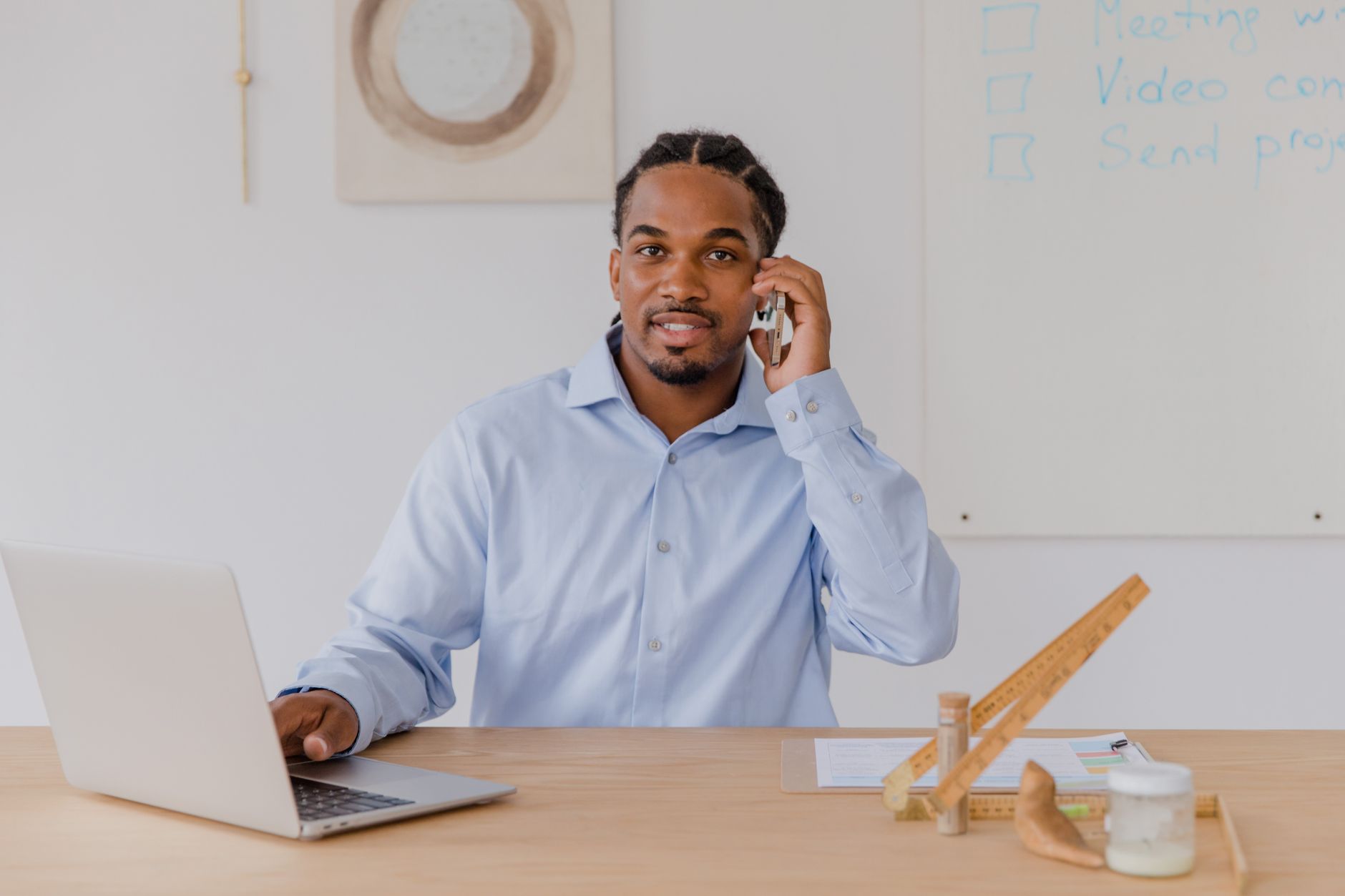 Jeune homme à son bureau, smartphone à la main, travaillant sur son ordinateur portable, souriant.