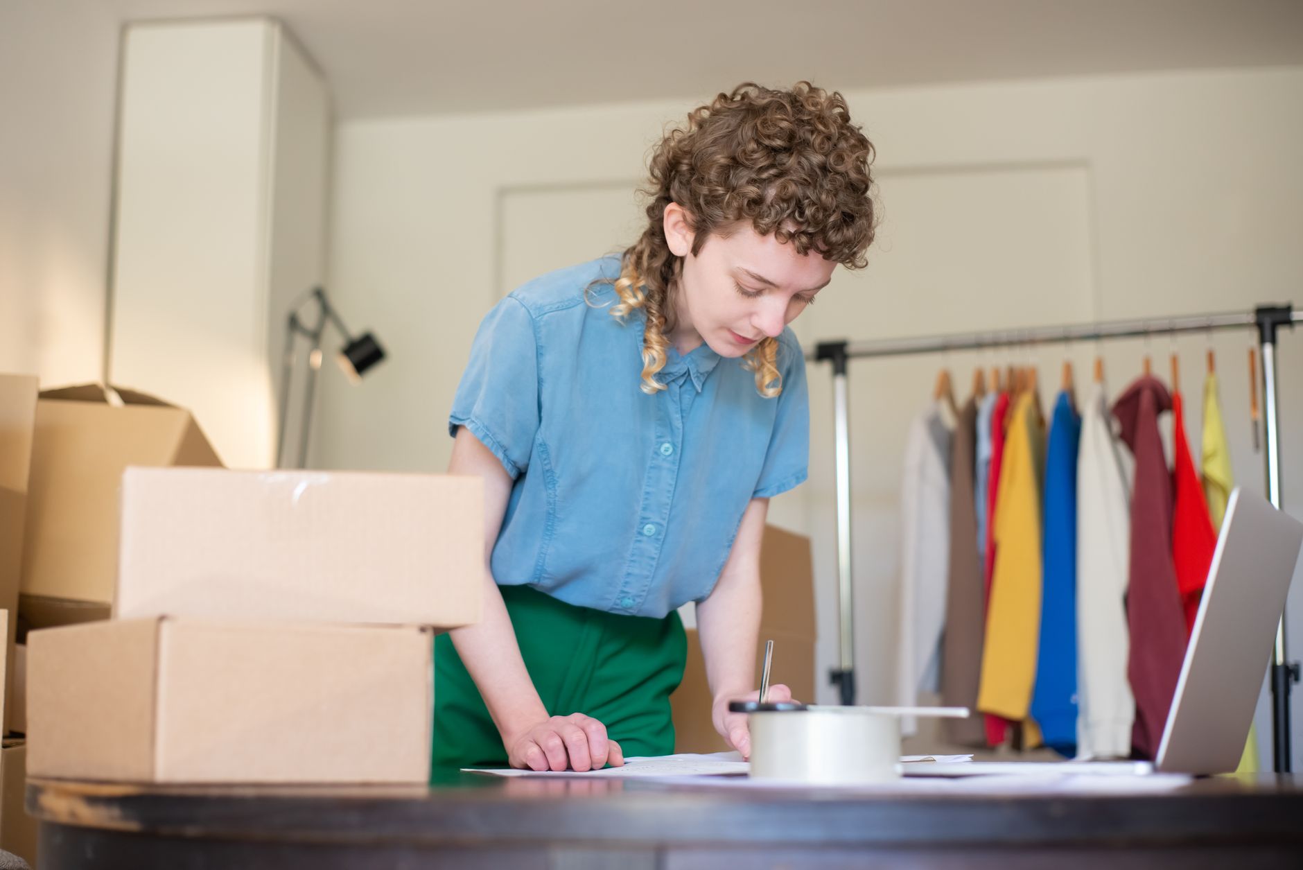 Femme préparant des commandes de sa boutique en ligne à domicile, entourée de cartons et d'un portant à vêtements.