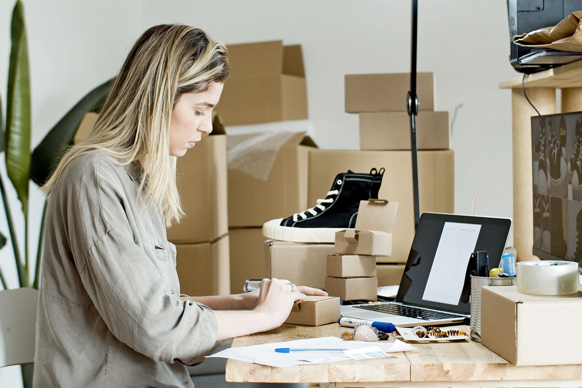Femme préparant des cartons pour sa boutique en ligne, entourée de matériel d'emballage et d'un ordinateur portable.